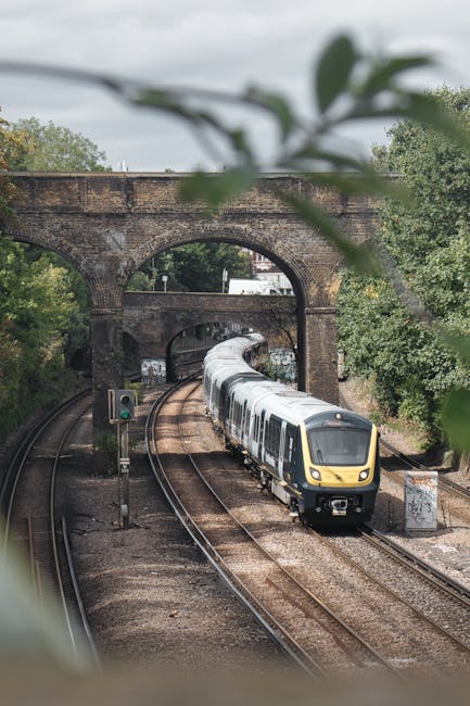 A modern passenger train with a yellow and grey front is traveling along curved railway tracks beneath a historic brick railway bridge, which features multiple arches supporting an elevated structure. Foreground leaves are partially blurred, framing the scene, while dense green trees and shrubs line the sides of the tracks, adding natural scenery to the setting. The scene is captured during daytime with overcast lighting, casting a soft, diffuse light over the train and surrounding environment. The tracks are laid on gravel beds, and signal poles with green lights are positioned along the line, indicating operational status. The presence of the railway bridge and passing train suggests an active rail corridor, which could relate to local independent or private transportation services, typically associated with alternative freight or passenger rail operations, aligning subtly with various waste management or logistic routes in urban or suburban areas such as Putney. Rubbish Collection Putney's service atmosphere surrounds this scene through the imagery's depiction of infrastructure and transit, indirectly supporting efficient transport and logistics tied to waste disposal and collection efforts in the region.