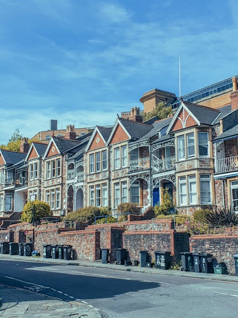 A row of Victorian-style terraced houses with ornate metalwork balconies and large bay windows, situated along a residential street with brick walls and front gardens that include well-maintained shrubs and small trees. The houses feature a combination of light-colored painted woodwork and red brick facades, with gabled roofs and decorative trim. At street level, there are multiple black wheelie bins lined up along the pavement, indicating waste collection activity, possibly handled by an independent rubbish removal service like Rubbish Collection Putney. The scene is illuminated by natural daylight under a clear blue sky with some wispy clouds, emphasizing the urban residential environment often associated with private waste disposal and on-site clearance options.