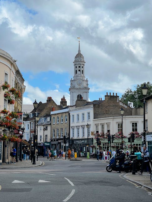 A busy street scene in a historic town center with a prominent white clock tower topped by a golden weather vane in the background. The street is lined with multi-story buildings featuring a mix of traditional and modern architectural styles, with some facades adorned with hanging flower baskets and decorative railings. Pedestrians are walking along the sidewalks, some engaging in conversation, while others browse shop windows or wait at crossings. On the right side, a police motorcycle is parked near a lamppost, and a few individuals are gathered nearby. The lower part of the image shows a paved road with marked lanes and a slight curve, with vehicles including a scooter and a small car, suggesting active traffic flow. Soft natural lighting filters through partially cloudy skies, creating a lively yet calm atmosphere typical of a central shopping and social area where private waste collection services, such as Rubbish Collection Putney, could facilitate efficient rubbish removal for local businesses and residents, supporting alternative waste management solutions beyond city council-operated services.