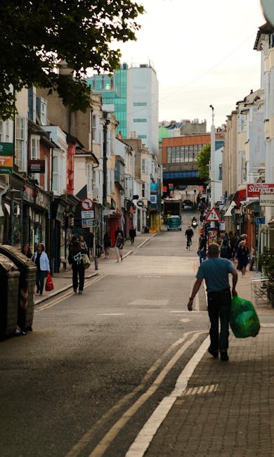 A busy urban street scene on Putney High Street, showing a mix of low-rise commercial and residential buildings on both sides with storefronts and signage, some featuring awnings and shop windows. The street is paved and slightly inclined, with pedestrians walking along the sidewalks and crossing at a marked pedestrian crossing station. In the foreground, a man dressed in a blue shirt and dark trousers is walking away from the camera, carrying a green bag of rubbish, indicating a private waste collection or alternative rubbish removal activity. The background features taller modern buildings with glass facades and a clear sky above, suggesting a typical day in a vibrant neighbourhood. The environment appears clean and well maintained, with a few vehicles parked along the roadside, highlighting the area's active commercial and residential character. The scene ostensibly relates to services like Rubbish Collection Putney providing on-site clearance or private disposal solutions for local households and businesses.